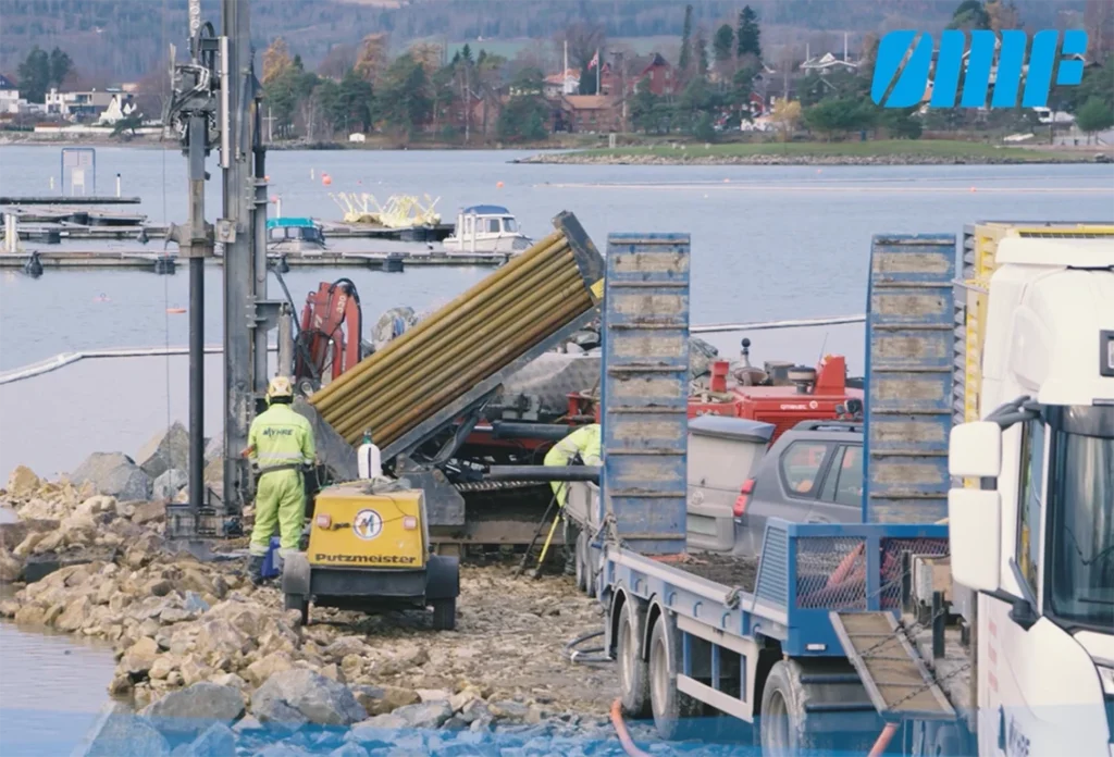 Arbeidere i varselklær bruker inklinometre mens de arbeider med tunge maskiner på en steinete byggeplass ved et vann, med båter og bygninger synlige i bakgrunnen.