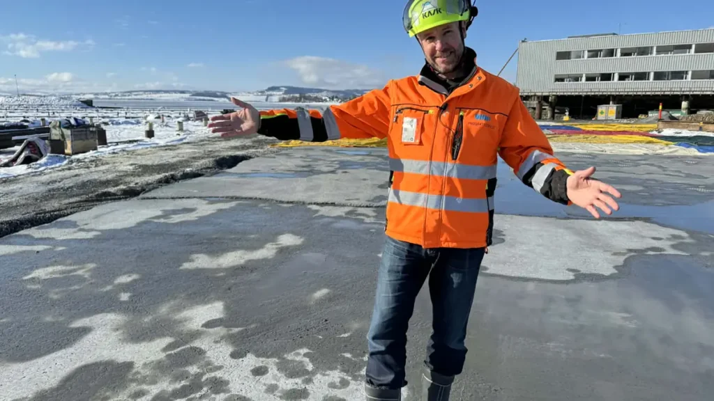 En anleggsarbeider i oransje vernetrøye og hjelm står med utstrakte armer på en delvis islagt flate, med 30 000 kubikk fyllt opp i Mjøsa og en snøkledd industribygning i bakgrunnen.