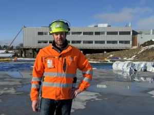 En anleggsarbeider iført oransje varseljakke og hjelm står utendørs på et vått, isete underlag med en stor industribygning og byggematerialer i bakgrunnen.