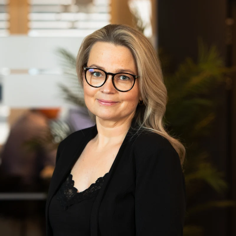 En kvinne med langt blondt hår og briller, iført svart topp og jakke, smiler mykt mens hun står innendørs med uskarp kontorbakgrunn og planter.