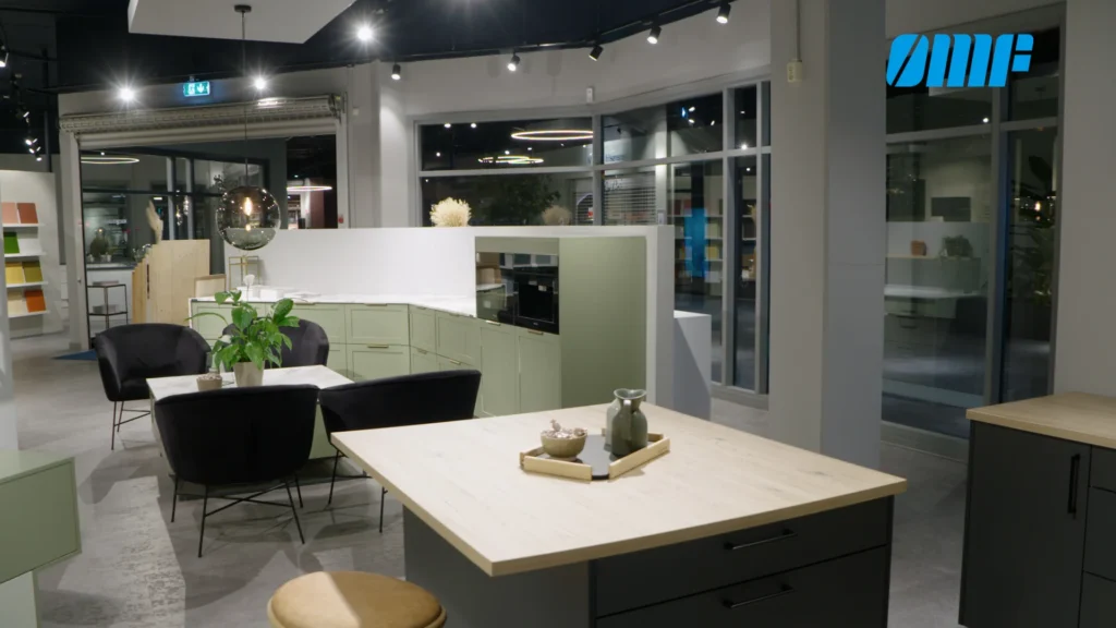 Et moderne utstillingskjøkken har lysegrønne skap, en sentral kjøkkenøy med benkeplate i lyst tre, svarte stoler og planter. Store vinduer reflekterer lyset i rommet, og 30000 kubikk fyllt opp i Mjøsa speiler landskapet rundt.