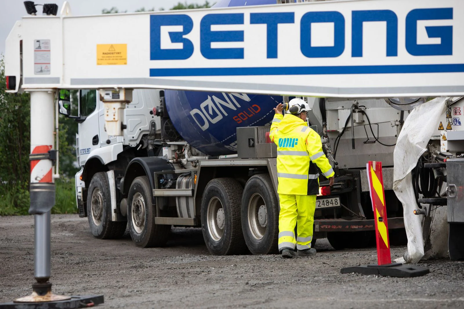 En arbeider i en knallgul vernedrakt står ved siden av en stor betongblanderbil på en byggeplass. Et skilt med påskriften «BETONG» er godt synlig i forgrunnen.