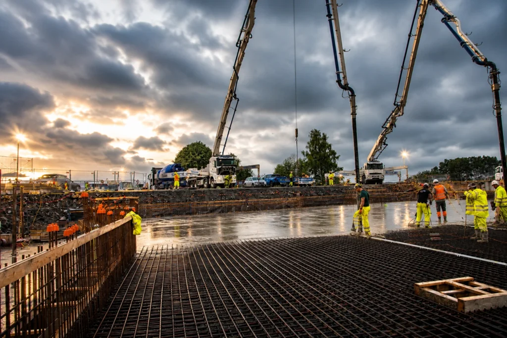 Byggarbeidere i verneutstyr er opptatt med å støpe en betongplate på en byggeplass på brygga i Hamar ved solnedgang, mens kraner og lastebiler tegner seg mot en dramatisk, overskyet himmel.