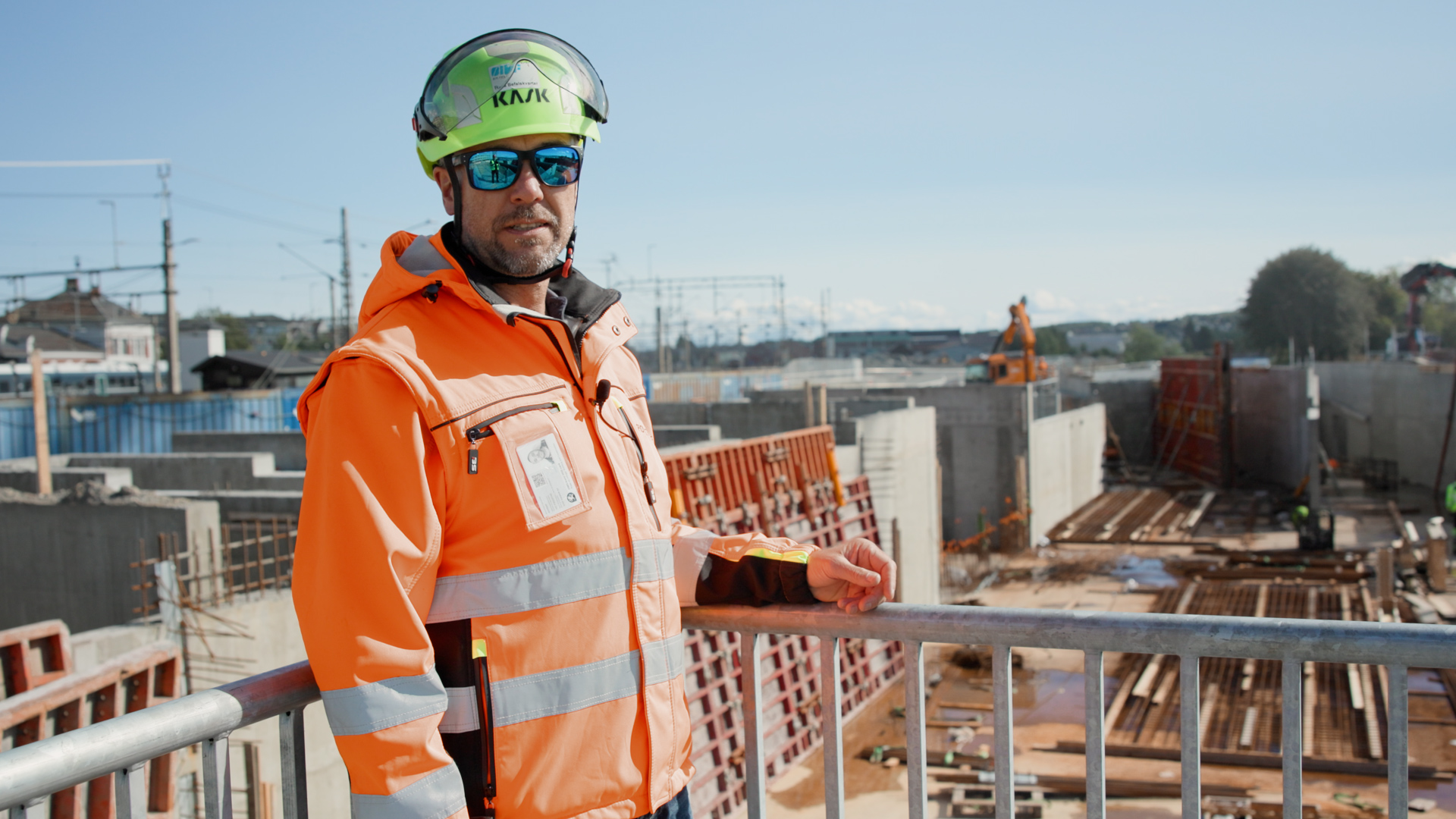 En bygningsarbeider i oransje sikkerhetsvest og hjelm står ved et rekkverk og ser ut over en byggeplass i full drift der støping av bunnplate pågår, med betongvegger og byggematerialer i bakgrunnen.